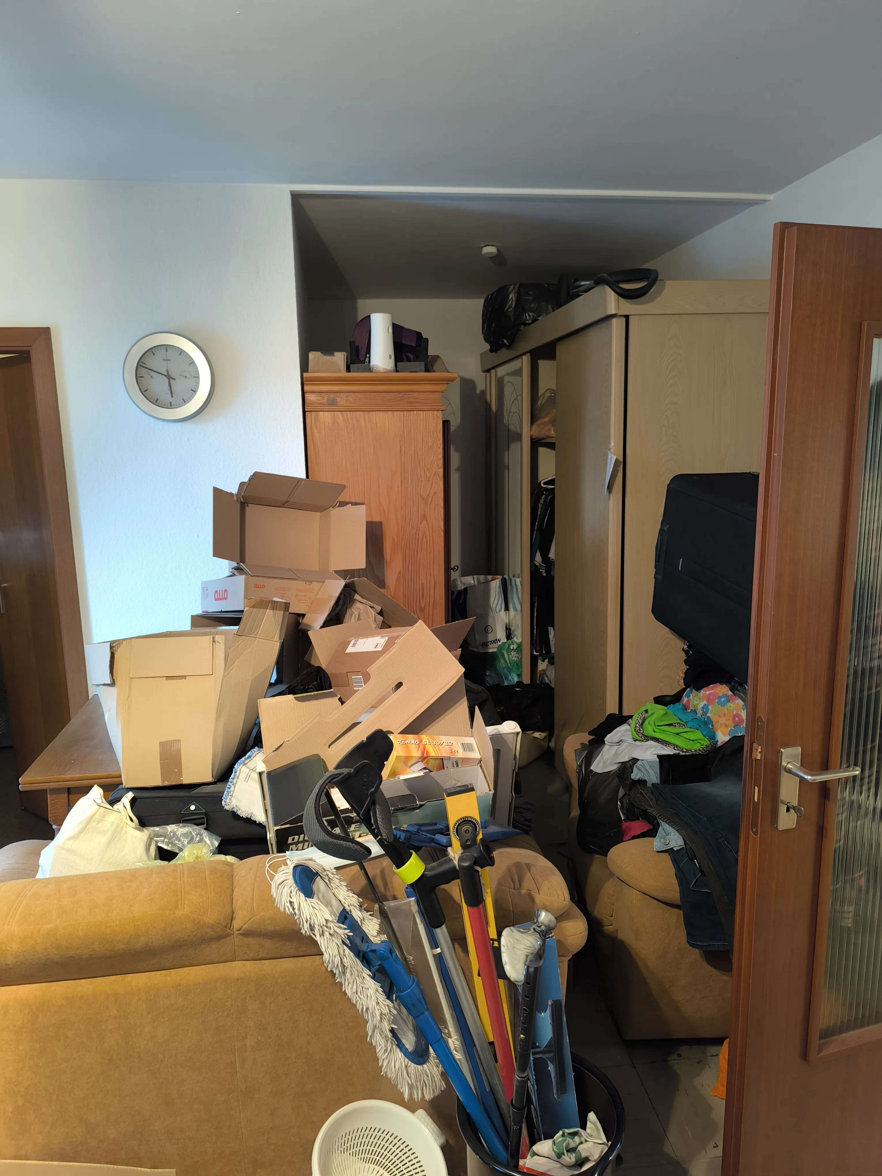 Unordentliches Wohnzimmer vor der Entrümpelung – voll mit Kartons, Kleidung, Möbeln und Reinigungsutensilien.